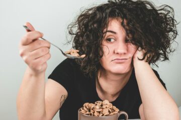 woman with messy hair wearing black crew-neck t-shirt holding spoon with cereals on top